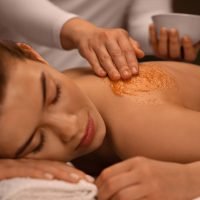 Closeup of sleeping young lady getting body treatment at spa, therapist applying scrub on woman shoulders